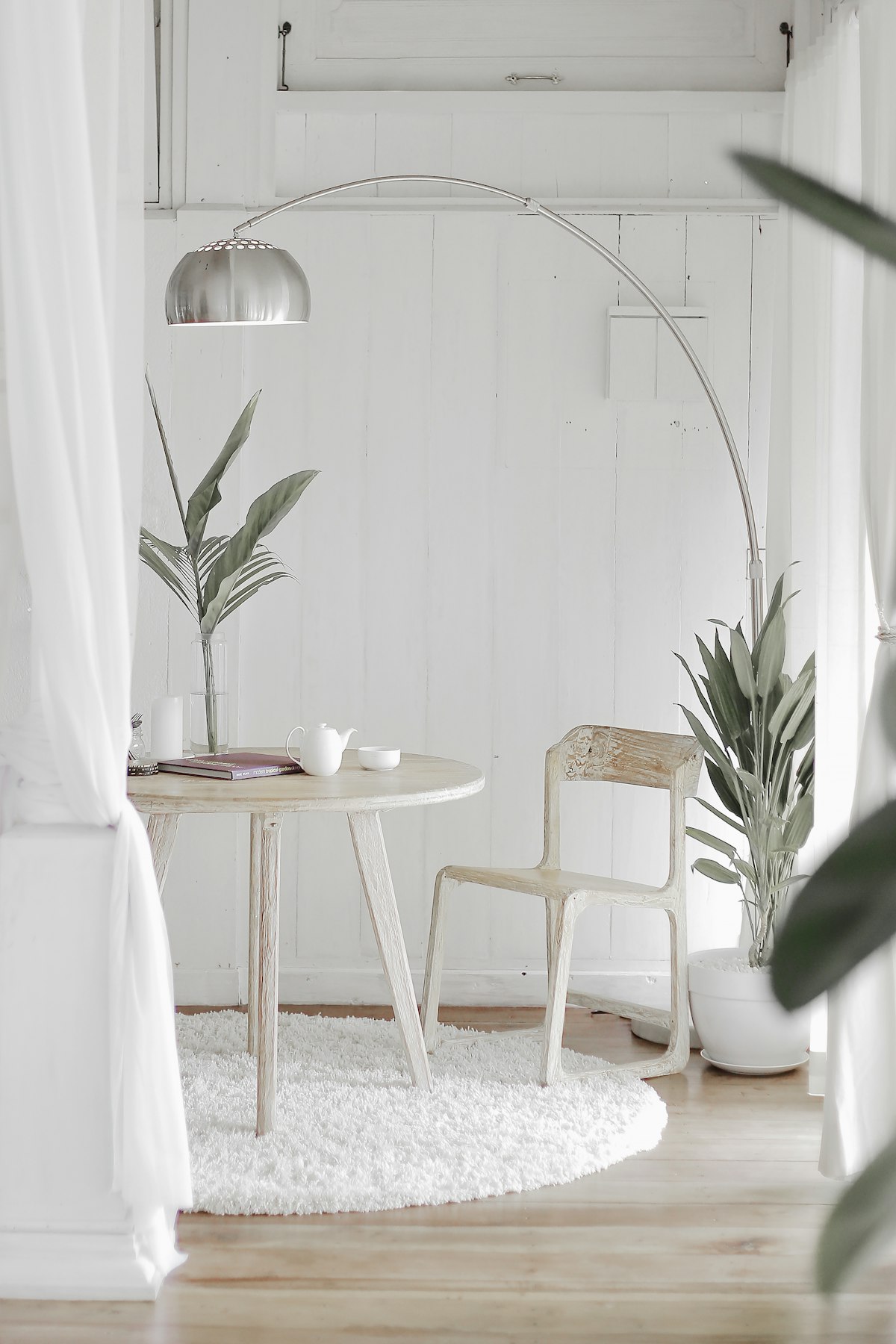 White curtains framing a wooden table, chair and arched floor lamp